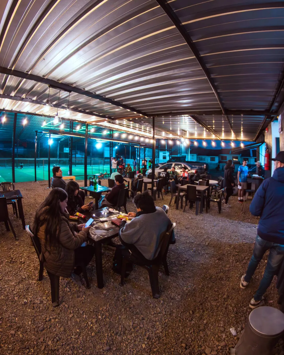 Área social llena de gente bajo las luces, con la cancha iluminada al fondo